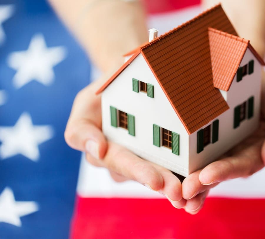 A hand holding a small model house with keys, against a U.S. flag